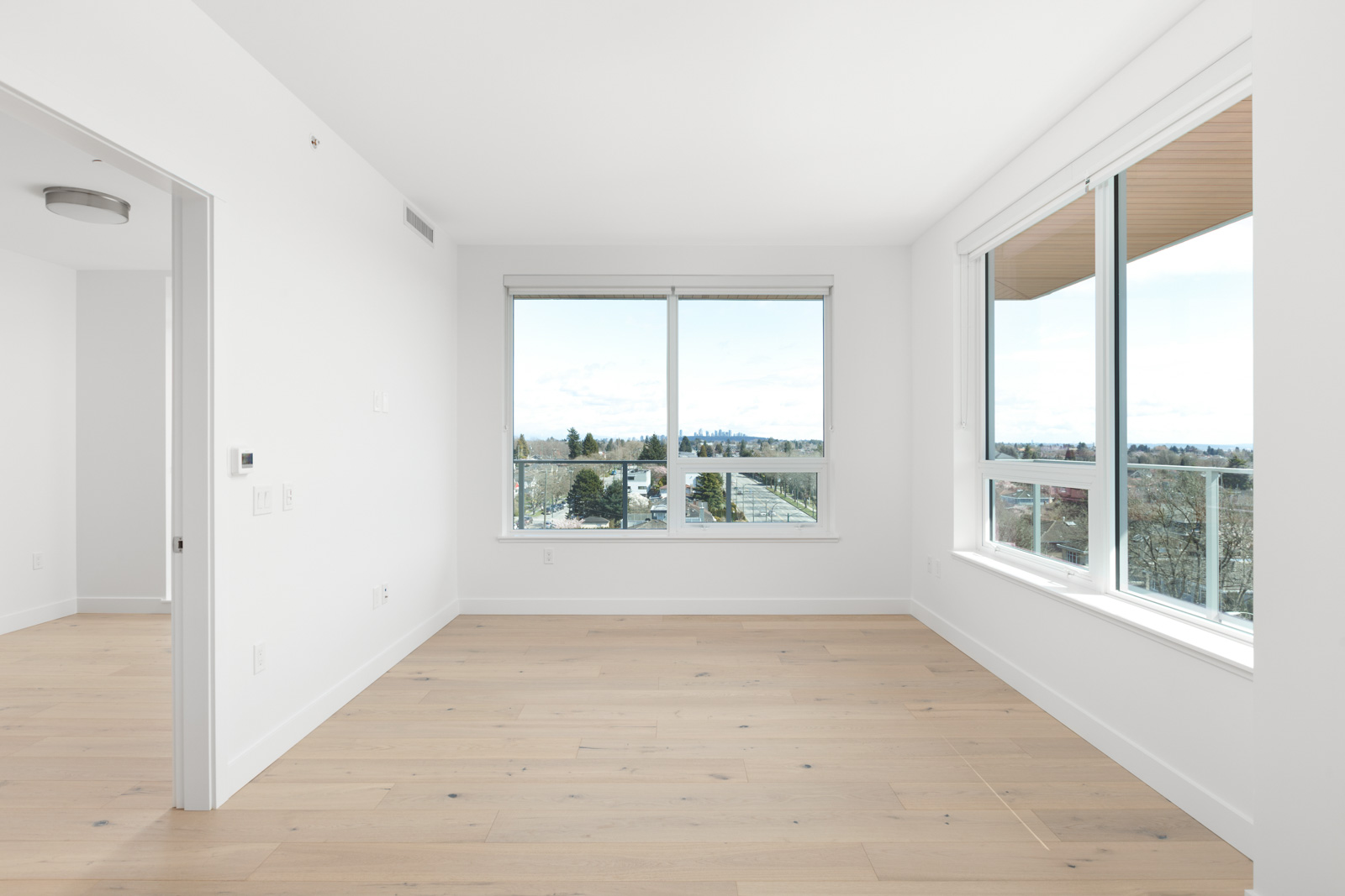 Empty, unfurnished room with large windows, light wood floors, and white walls, offering a view of trees and a distant city skyline.