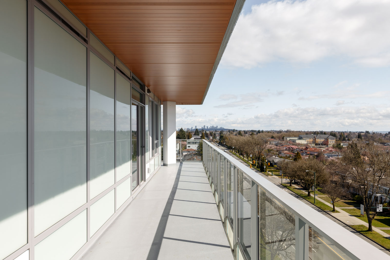 A modern apartment balcony with glass railing overlooks a residential neighborhood and distant city skyline under a partly cloudy sky.
