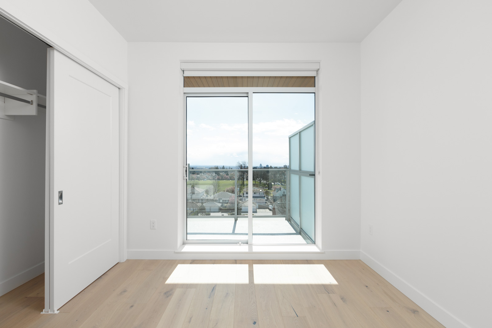 Empty, white-walled room with light wood floor, open closet door, and glass sliding doors leading to a balcony with a view of trees and sky.