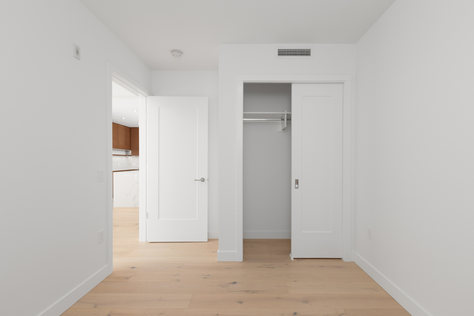 Empty white room with wooden floor, an open door leading to a kitchen, and a small closet with a sliding door and a hanging rod.