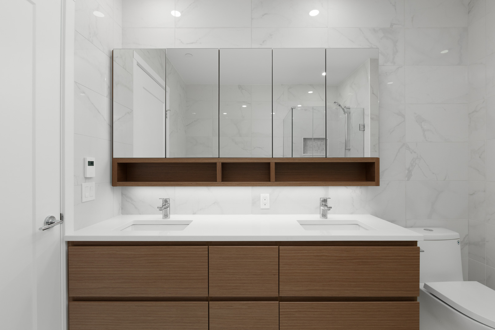 Modern bathroom with double sink vanity, wood cabinetry, large mirrored cabinets, white marble walls, and recessed lighting.