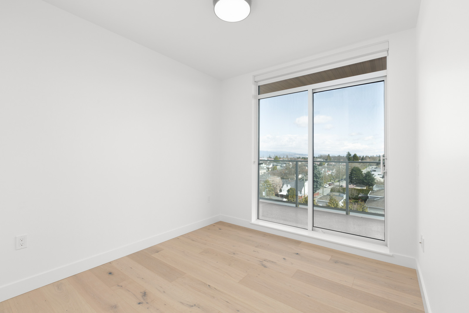 Empty room with light wood flooring, white walls, and a large window with sliding glass doors leading to a balcony overlooking a cityscape.