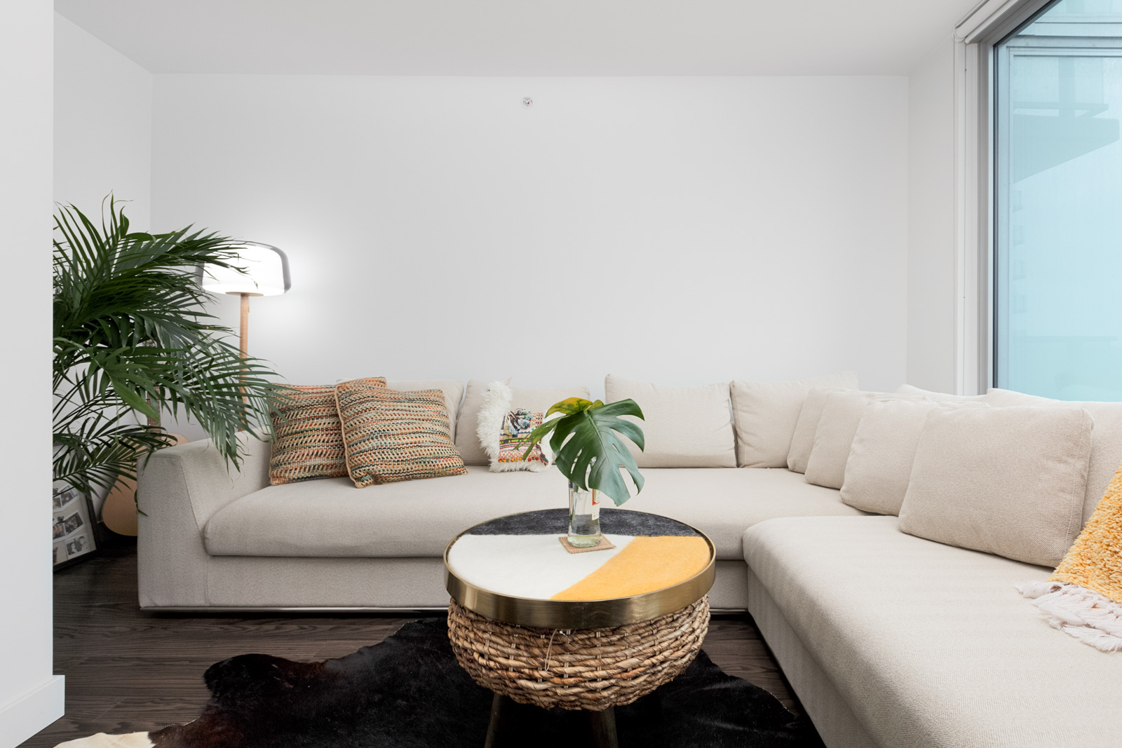 A modern living room with a light beige sectional sofa, patterned cushions, a round coffee table with a small plant, and a large green potted plant in the corner.