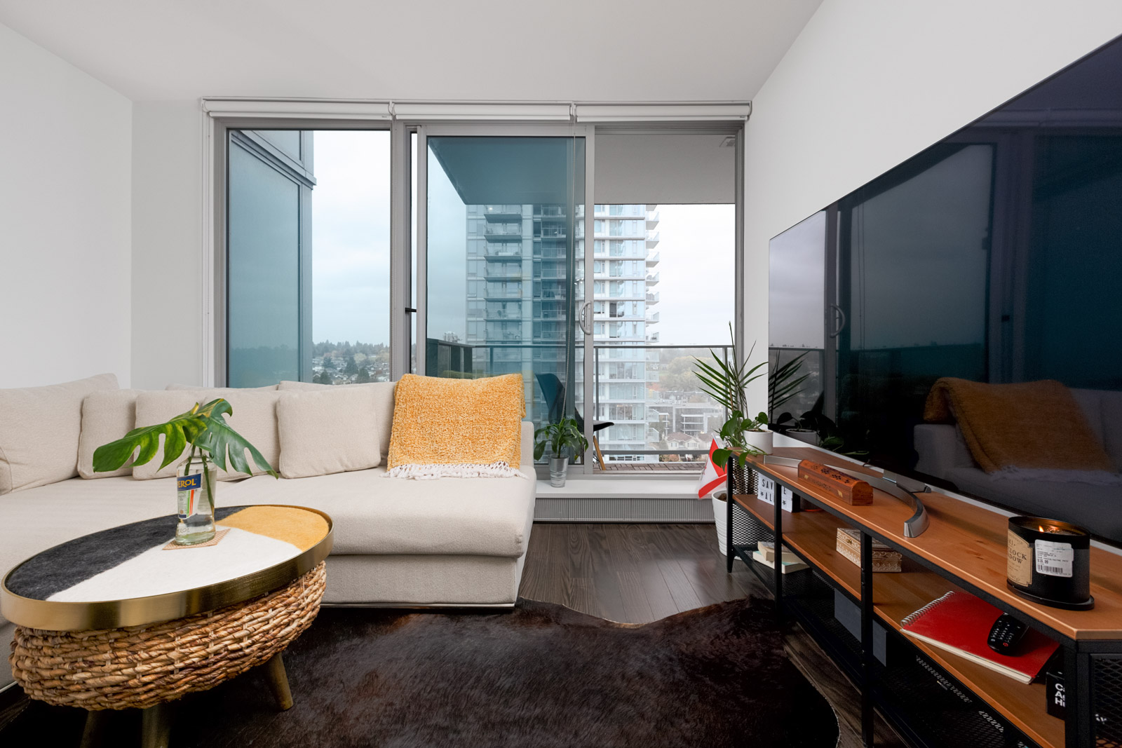 A modern living room with a beige sectional sofa, round coffee table, large TV, and a view of city buildings through floor-to-ceiling windows.