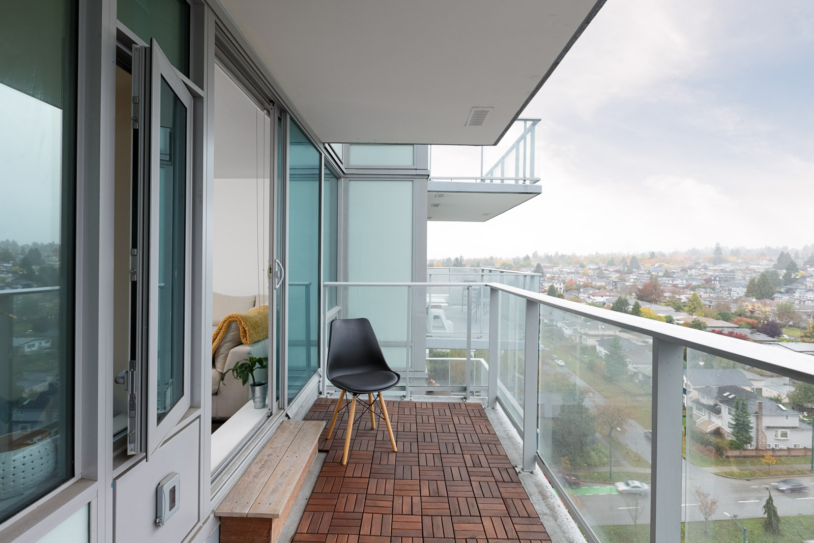 A modern apartment balcony with glass railings, wood tile flooring, a black chair, and a city view on a cloudy day.