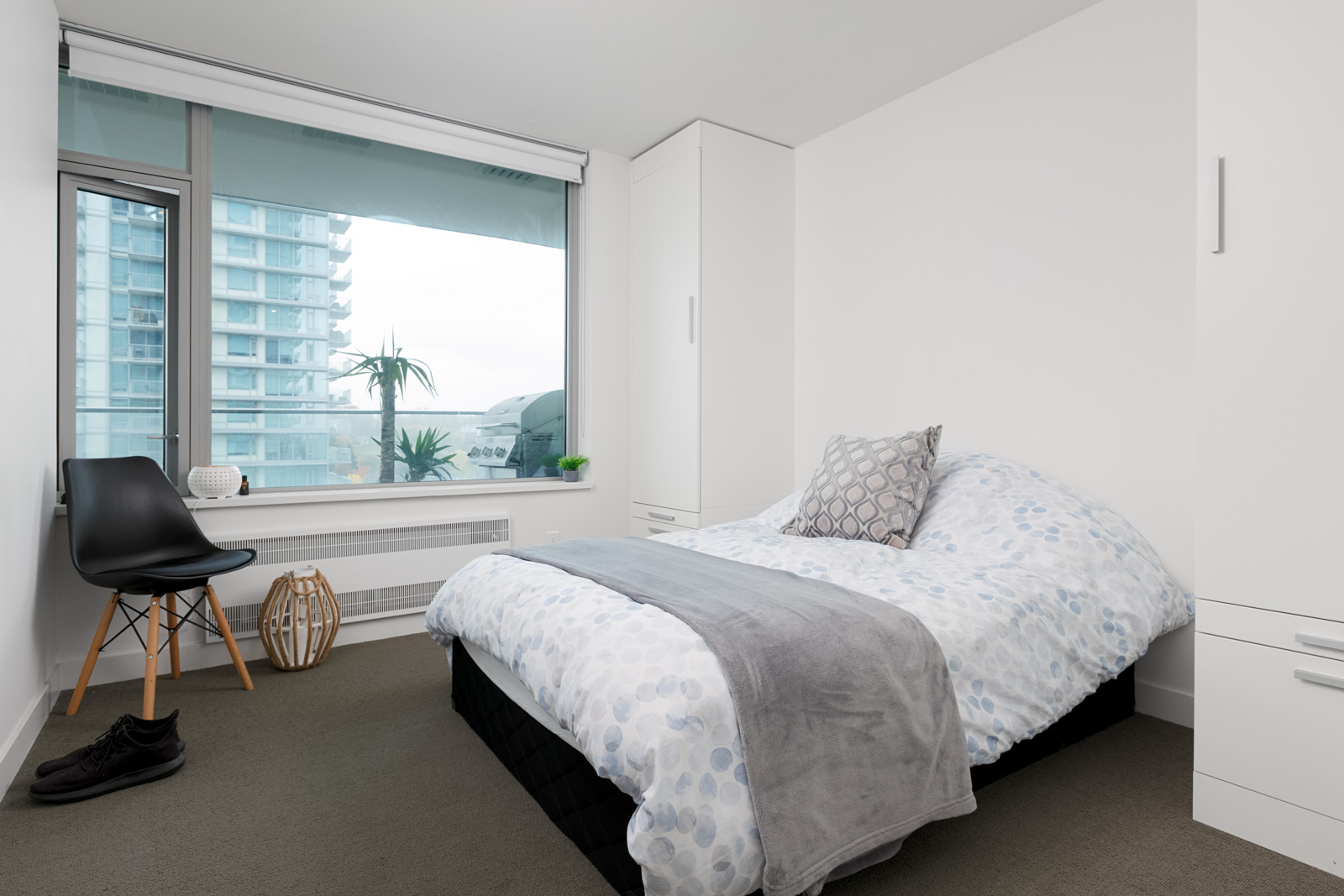 A modern bedroom with a neatly made bed, a black chair, a window with a city view, white walls, and built-in cabinetry.