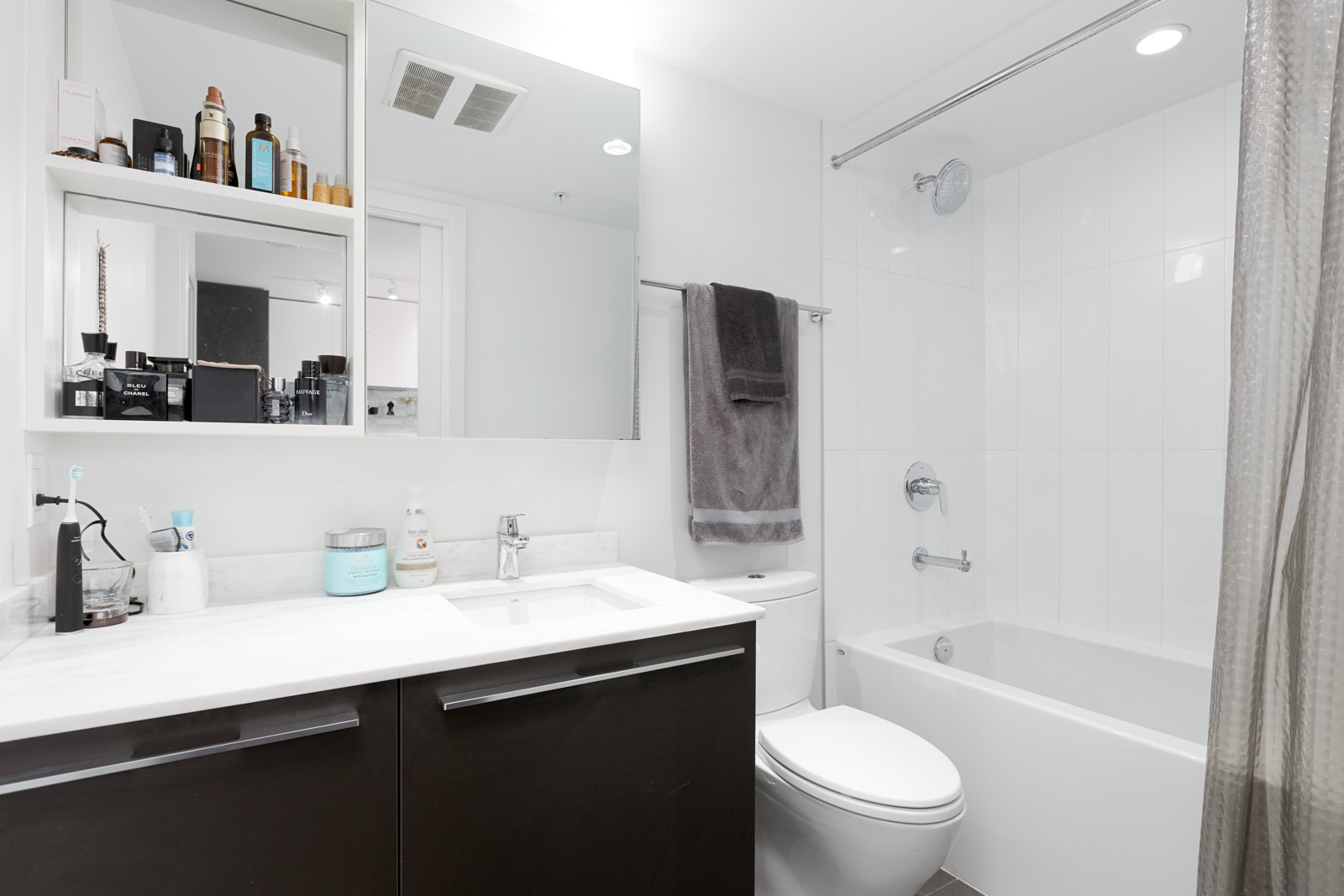 Modern bathroom with a sink, mirror, shelves with toiletries, towel on rack, toilet, and a bathtub with a shower and glass curtain.
