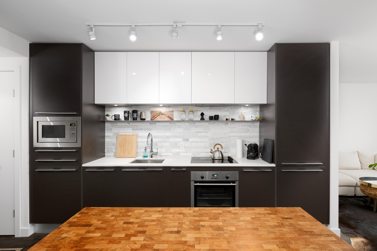 Modern kitchen with dark cabinets, white upper cupboards, built-in microwave, oven, sink, and open shelves, viewed from behind a wooden island countertop.