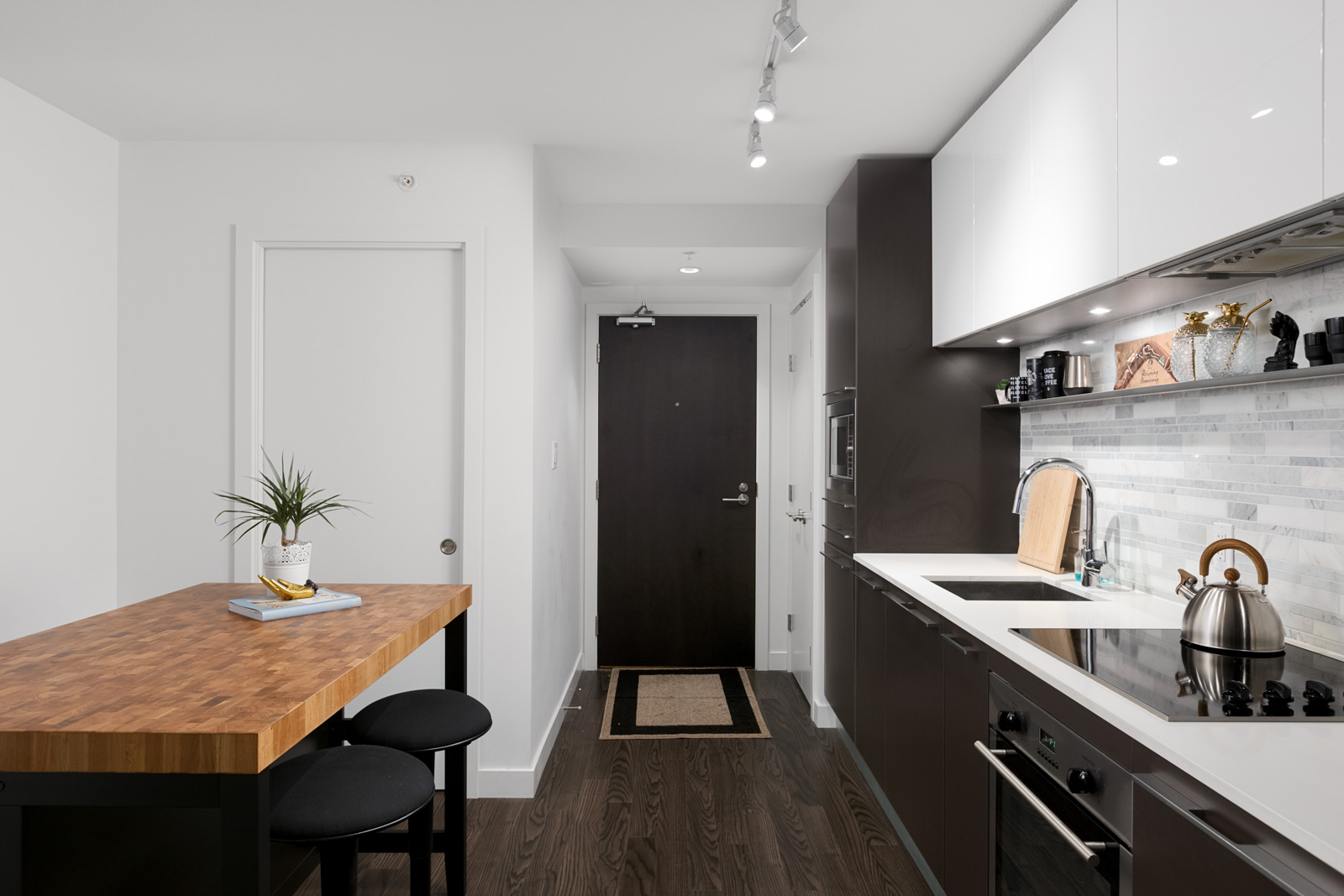 Modern kitchen with dark wood floors, a wooden island with two stools, white and dark cabinets, and a stainless steel stove and kettle. A door is visible at the end of the room.