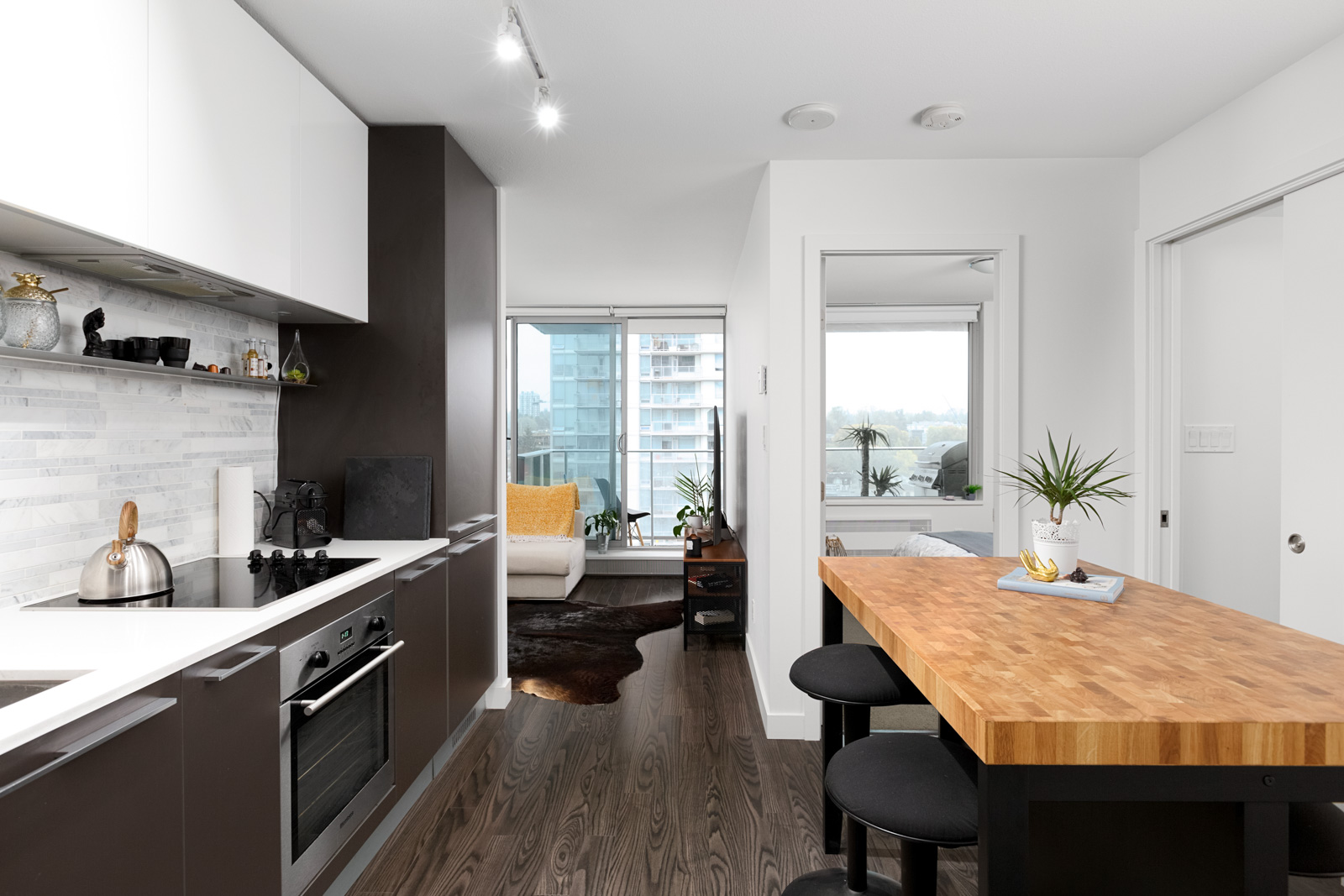 Modern kitchen with black and white cabinetry, wood countertops, bar stools, and an open view into a bright living area with large windows.
