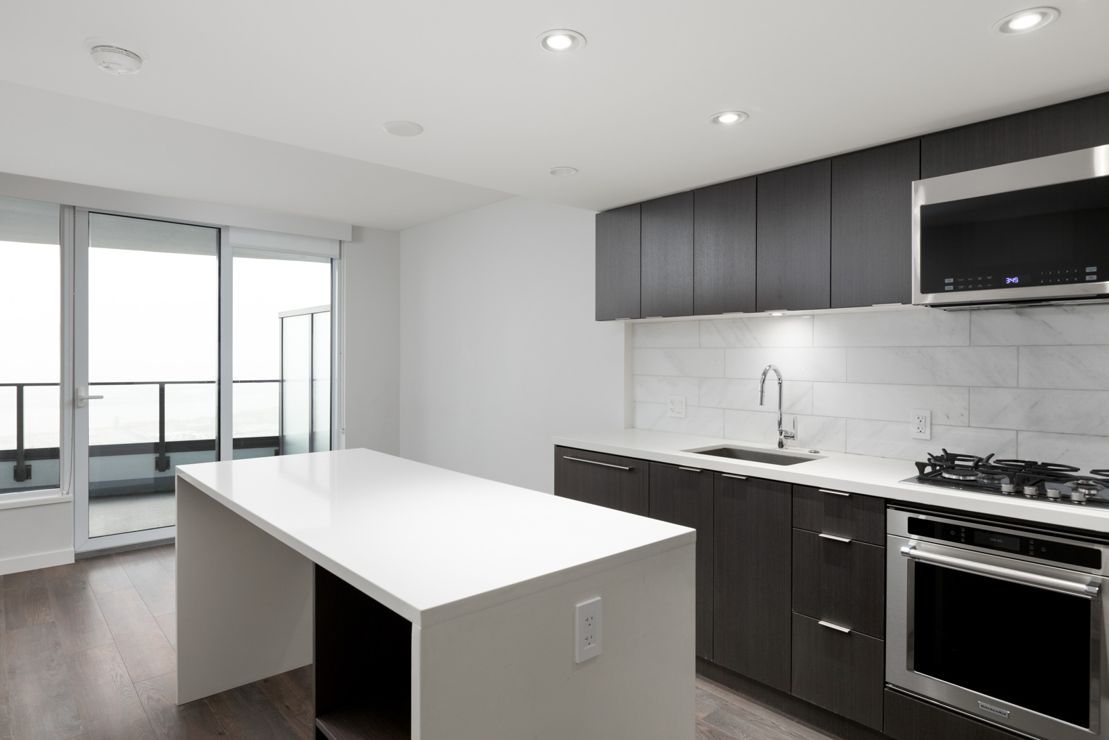 Modern kitchen with dark cabinets, white countertops, built-in appliances, and sliding glass doors leading to a balcony.