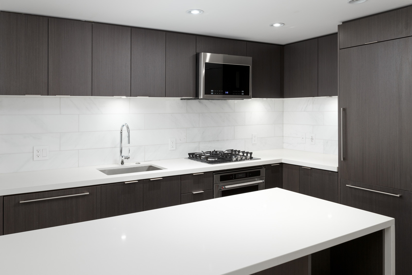 Modern kitchen with dark cabinets, white countertops, a built-in gas cooktop, oven, sink, and stainless steel microwave, with white tile backsplash and under-cabinet lighting.