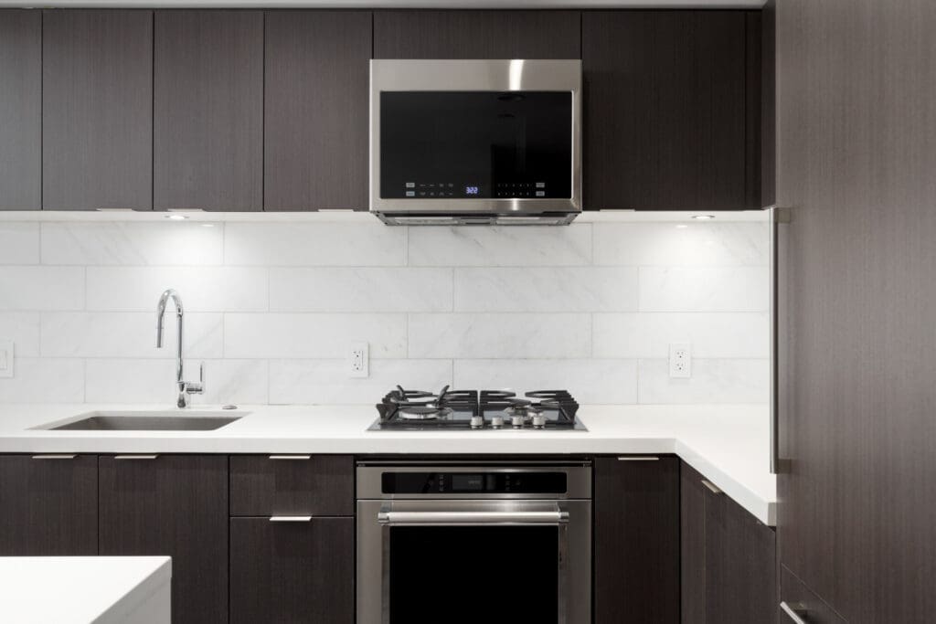 Modern kitchen with dark cabinets, white countertops, a built-in oven, gas stove, stainless steel range hood, and a sink with a gooseneck faucet against a tiled backsplash.