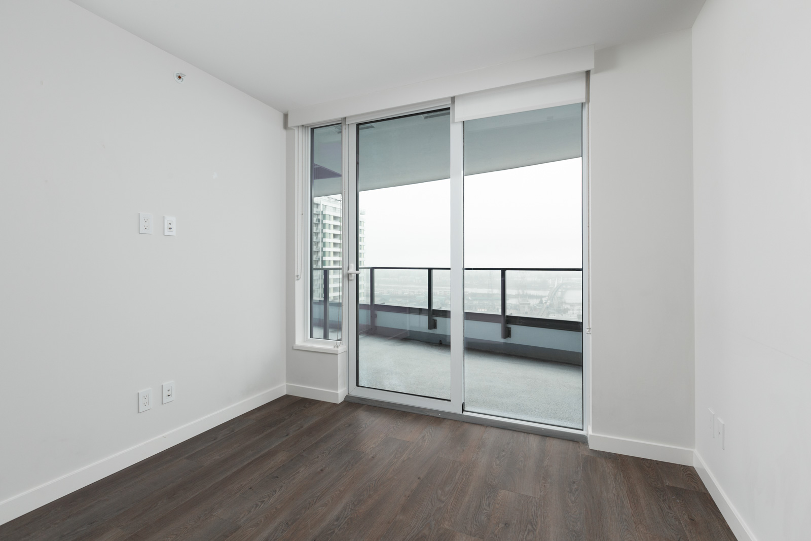 Unfurnished room with dark wood flooring, white walls, and a large glass door leading to a balcony overlooking city buildings.