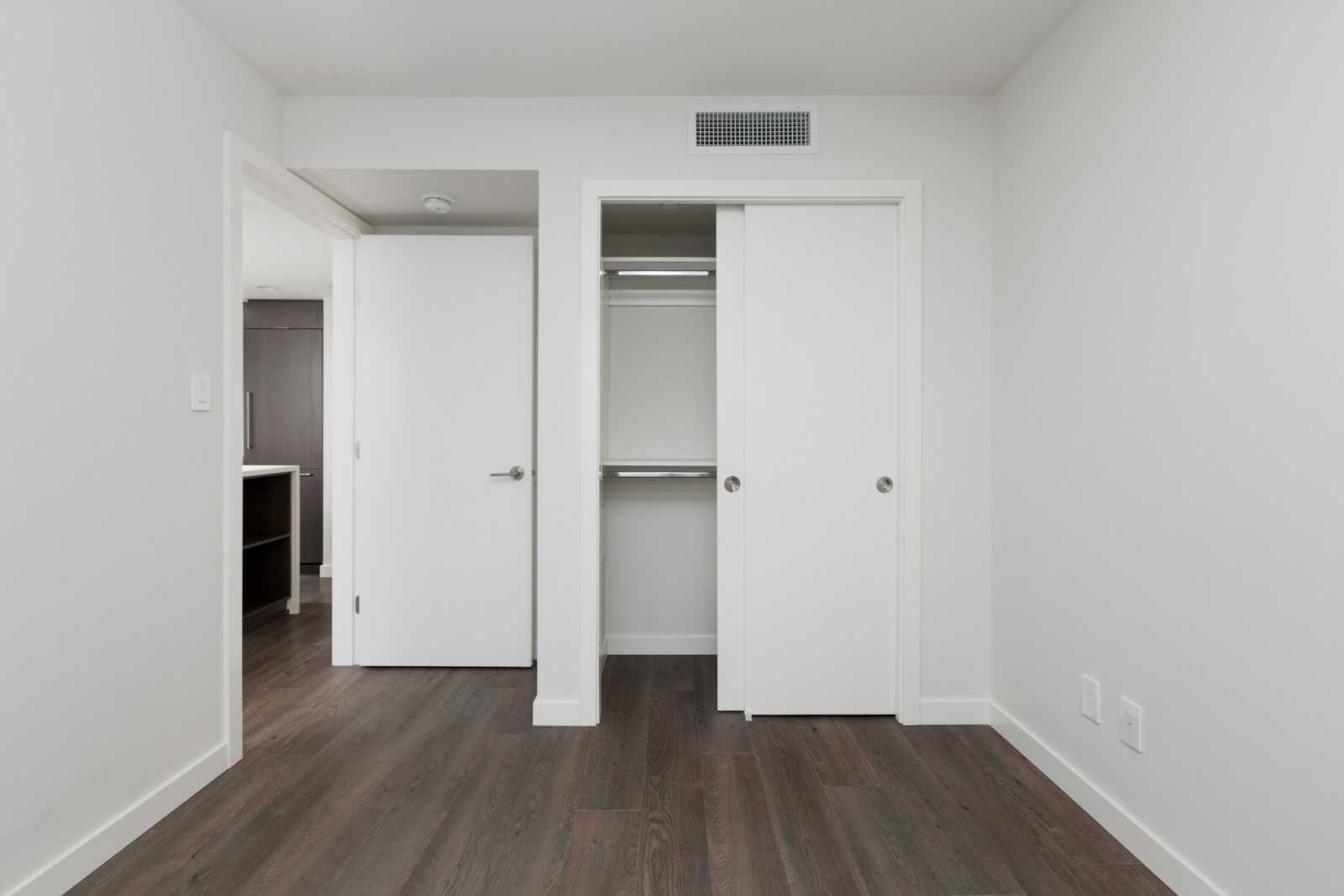 Empty room with white walls, dark wood flooring, an open closet with sliding doors, and an open door leading to another area.