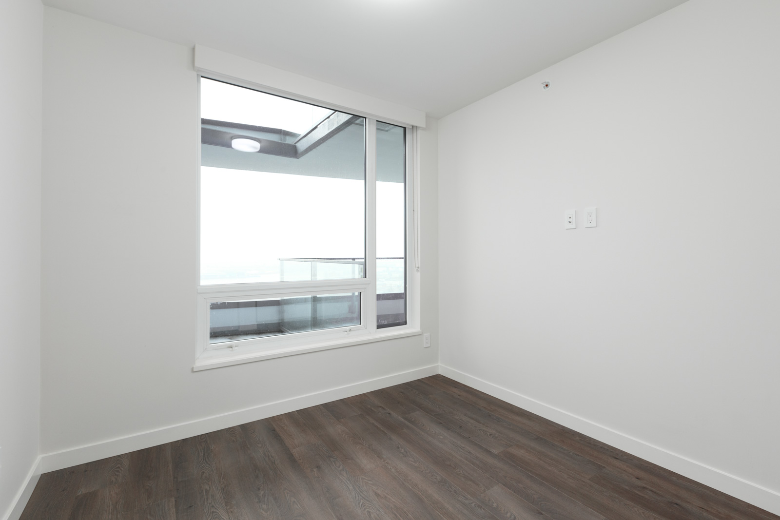 Empty room with white walls, dark wood flooring, and a large window overlooking a balcony with a glass railing.