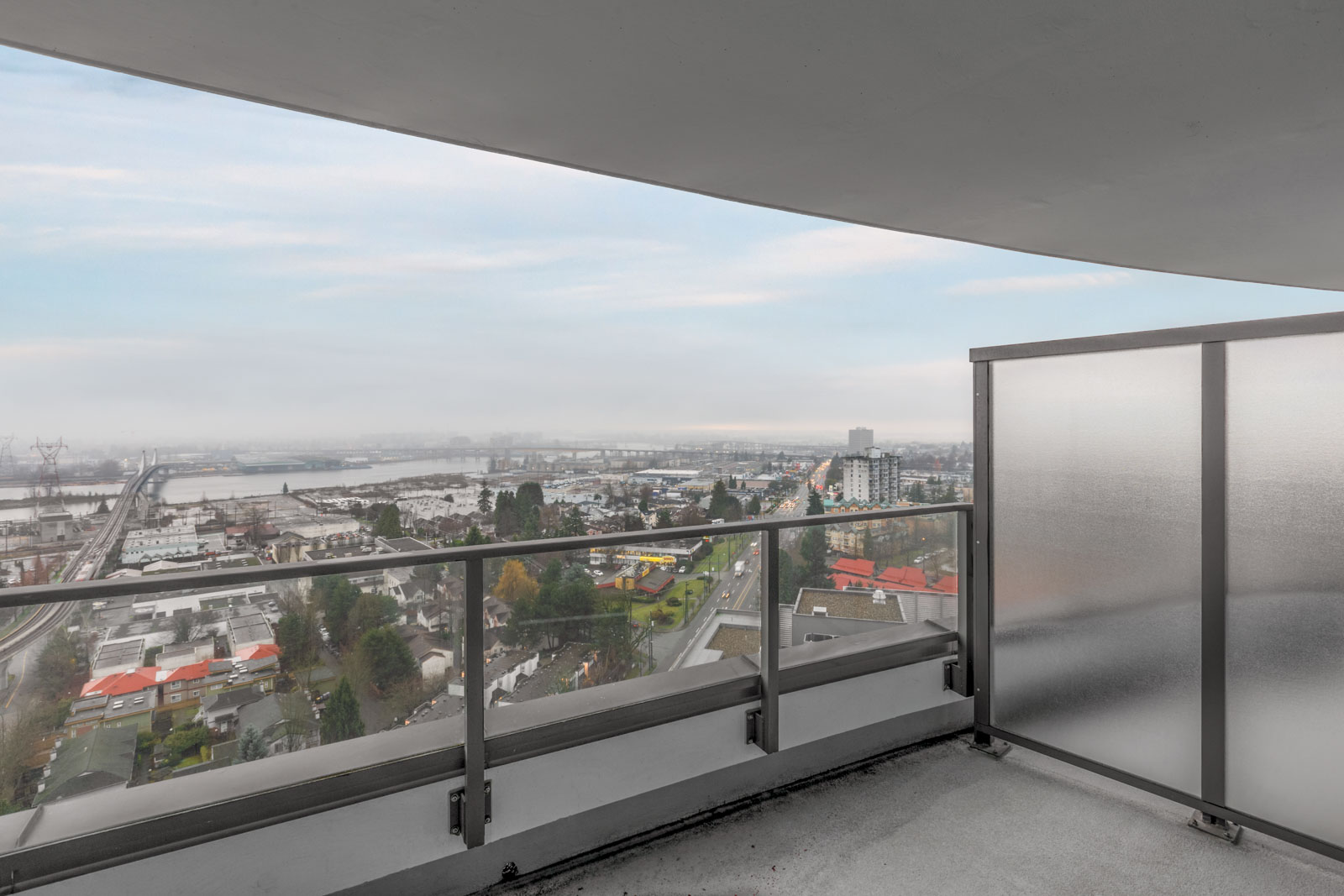 View from a high-rise balcony with glass railing, overlooking a cityscape, river, and distant industrial area under a cloudy sky.
