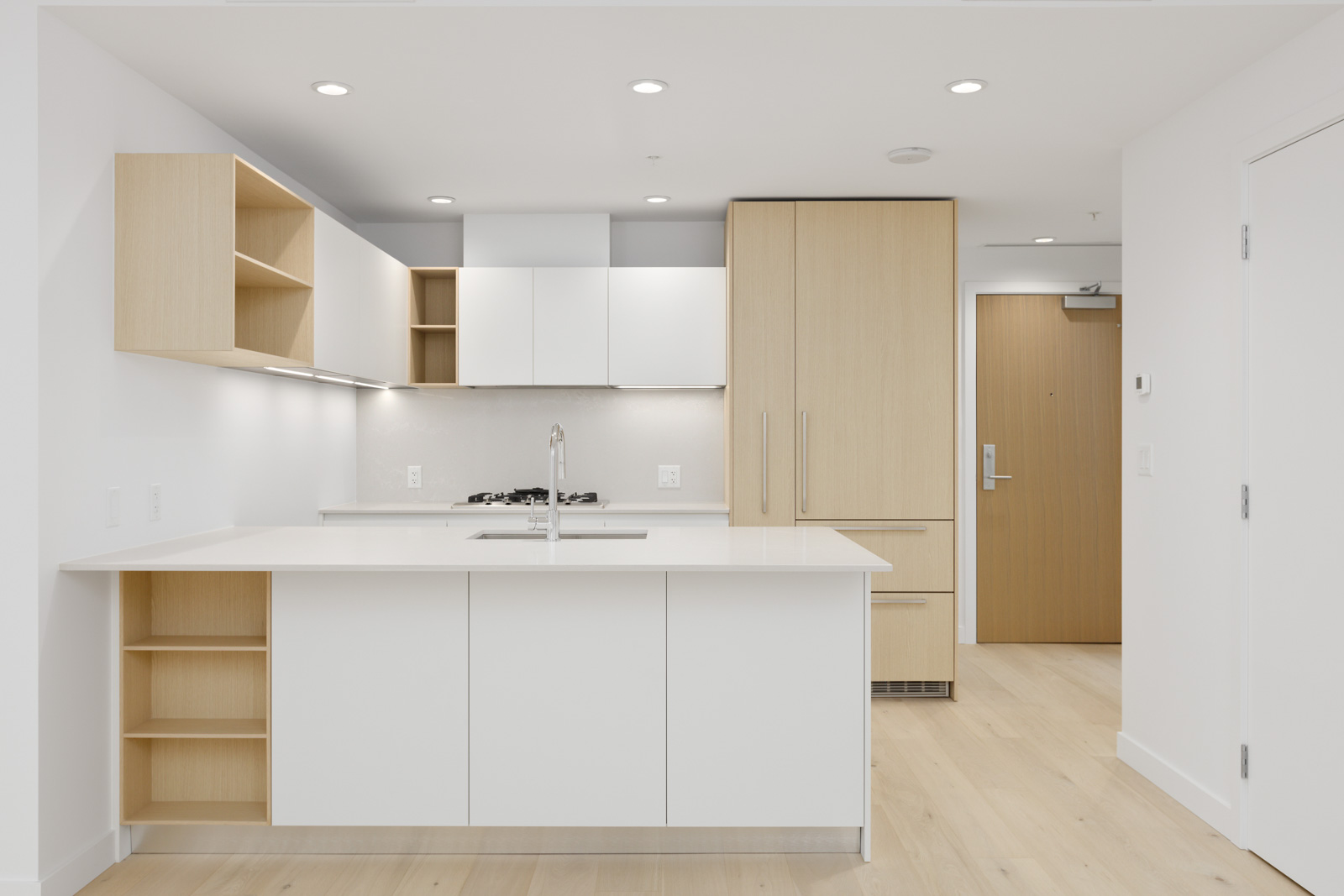 Modern kitchen with white cabinets, built-in appliances, light wood accents, open shelving, and an island sink under recessed lighting.