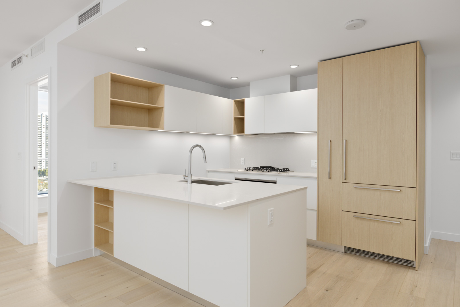 Modern kitchen with light wood and white cabinetry, built-in appliances, an island with a sink, and open shelves, featuring light-colored flooring and recessed lighting.
