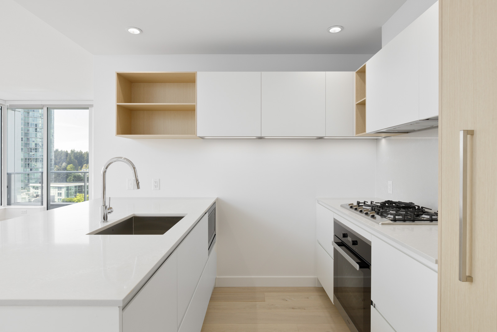 Modern white kitchen with built-in oven, gas cooktop, sink, open shelving, and large windows overlooking city buildings.