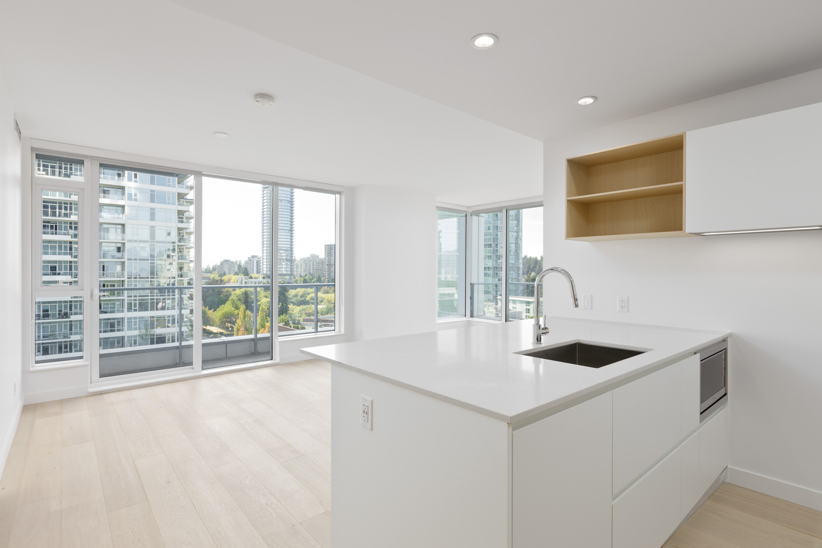 Modern, unfurnished apartment with large windows, light wood floors, white kitchen cabinets, and a sink island, overlooking city buildings and greenery.
