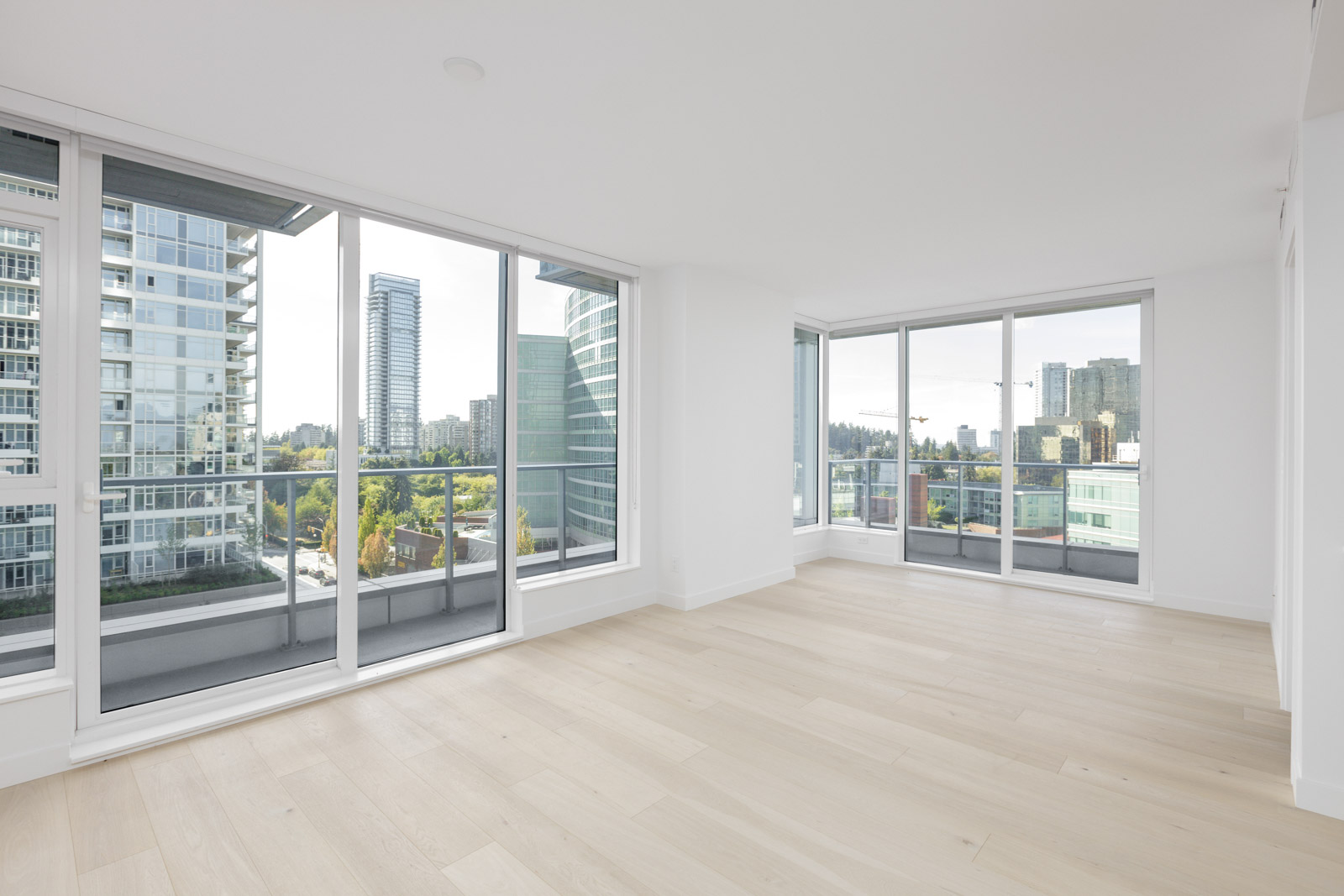 Empty modern apartment with light wood floors, large floor-to-ceiling windows, and a cityscape view from the balcony.