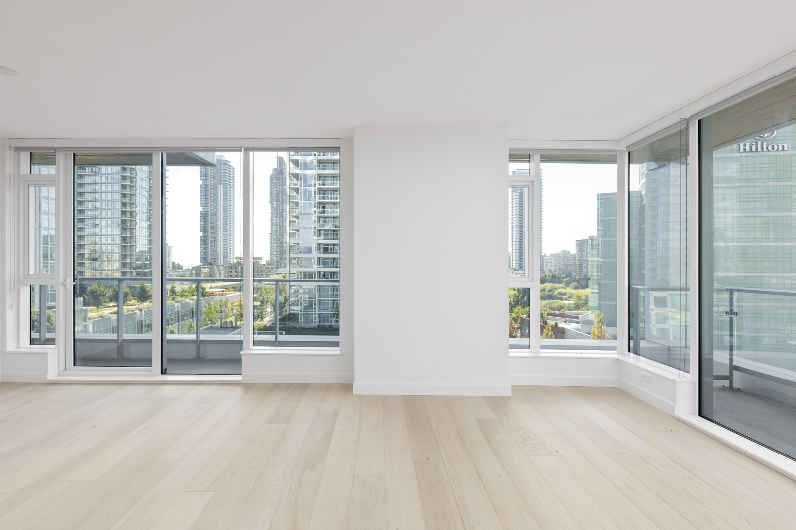 Unfurnished modern apartment with large windows, light wood floors, and a balcony overlooking a cityscape with high-rise buildings.
