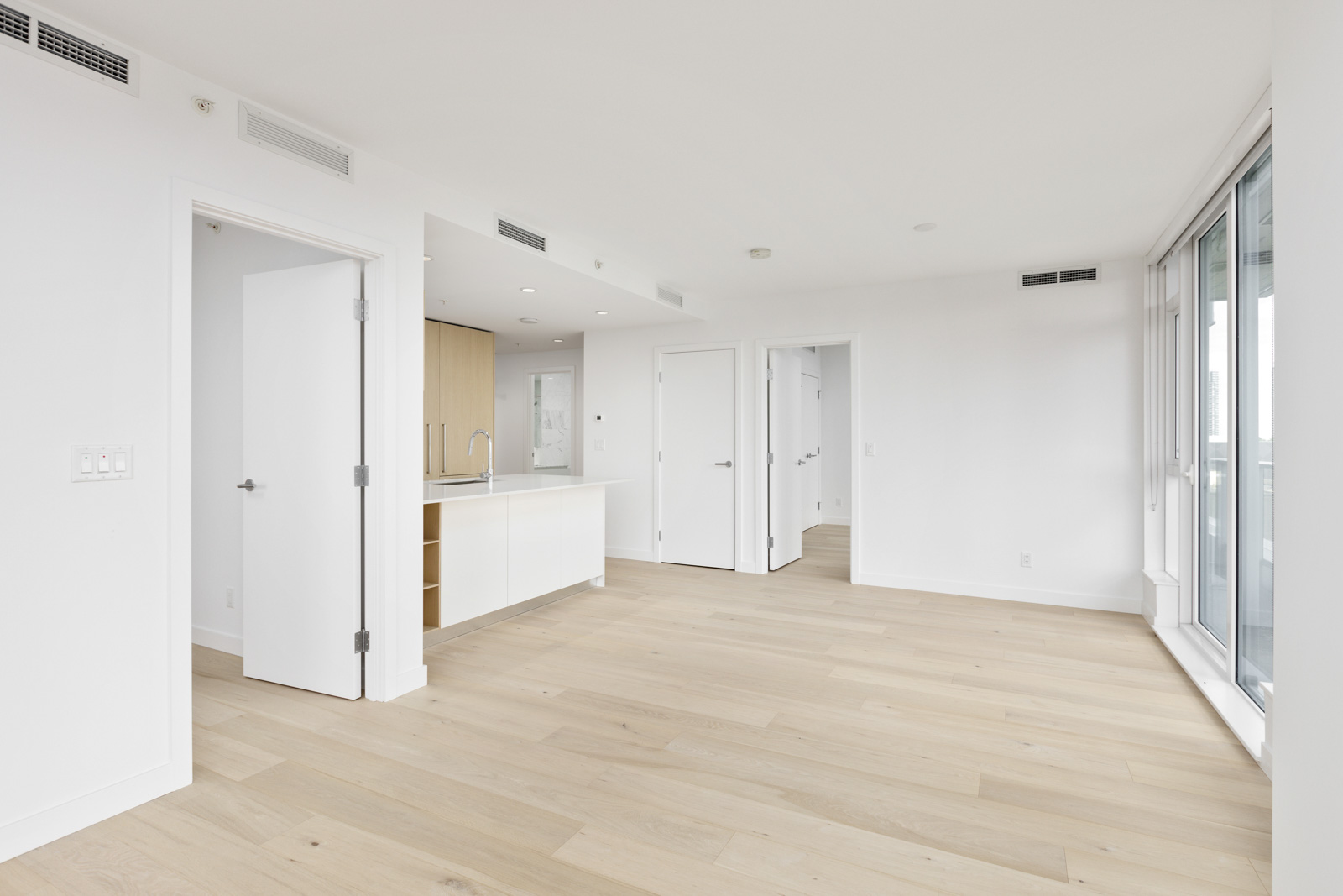 Empty modern apartment with light wood floors, white walls, open kitchen area, multiple doors, and large floor-to-ceiling windows.