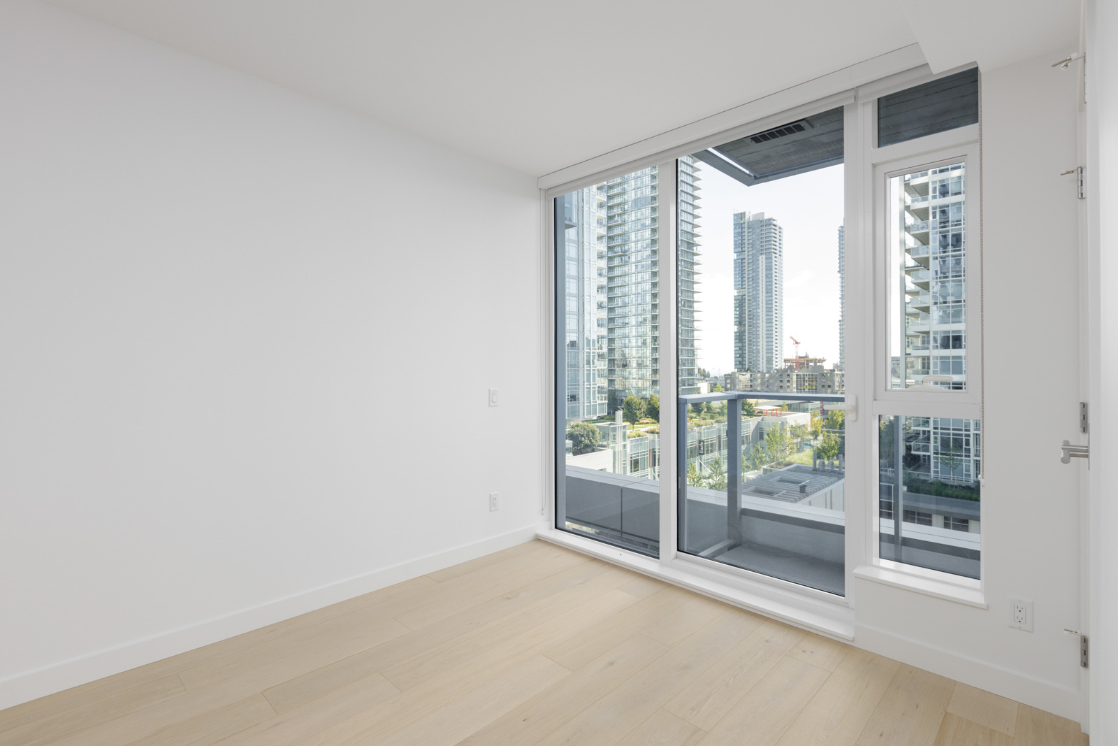 Unfurnished room with light wood floors, white walls, and large floor-to-ceiling windows leading to a balcony with views of modern city buildings.