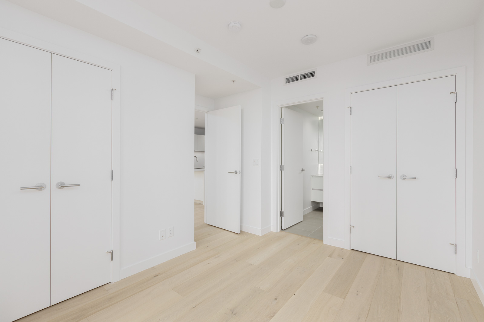A modern, empty room with light wood flooring, white walls, and several white doors, including a bathroom and closet doors, under bright natural light.
