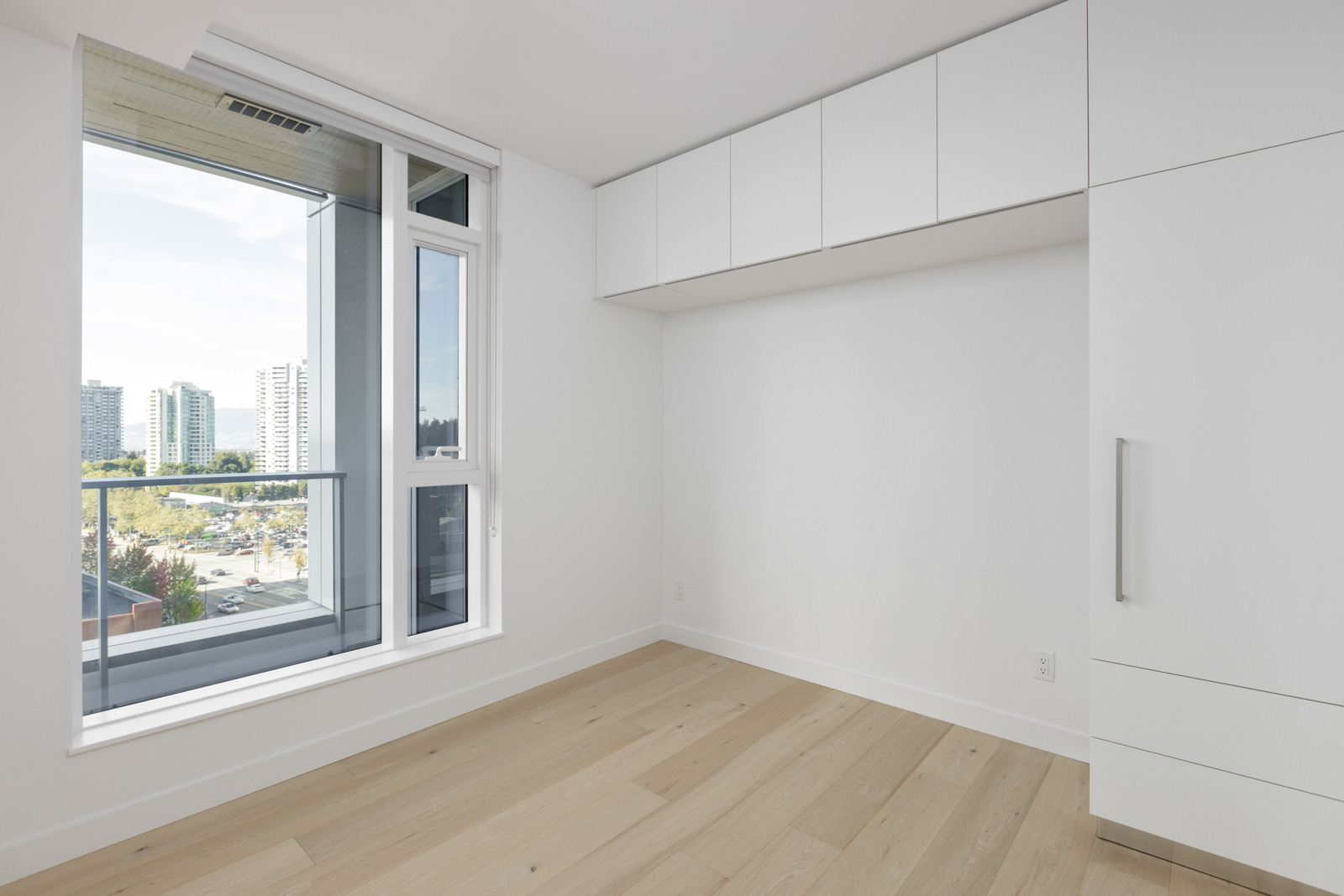 Empty modern room with light wood flooring, white walls, built-in cabinets, and a large window showing a city view with buildings and trees.