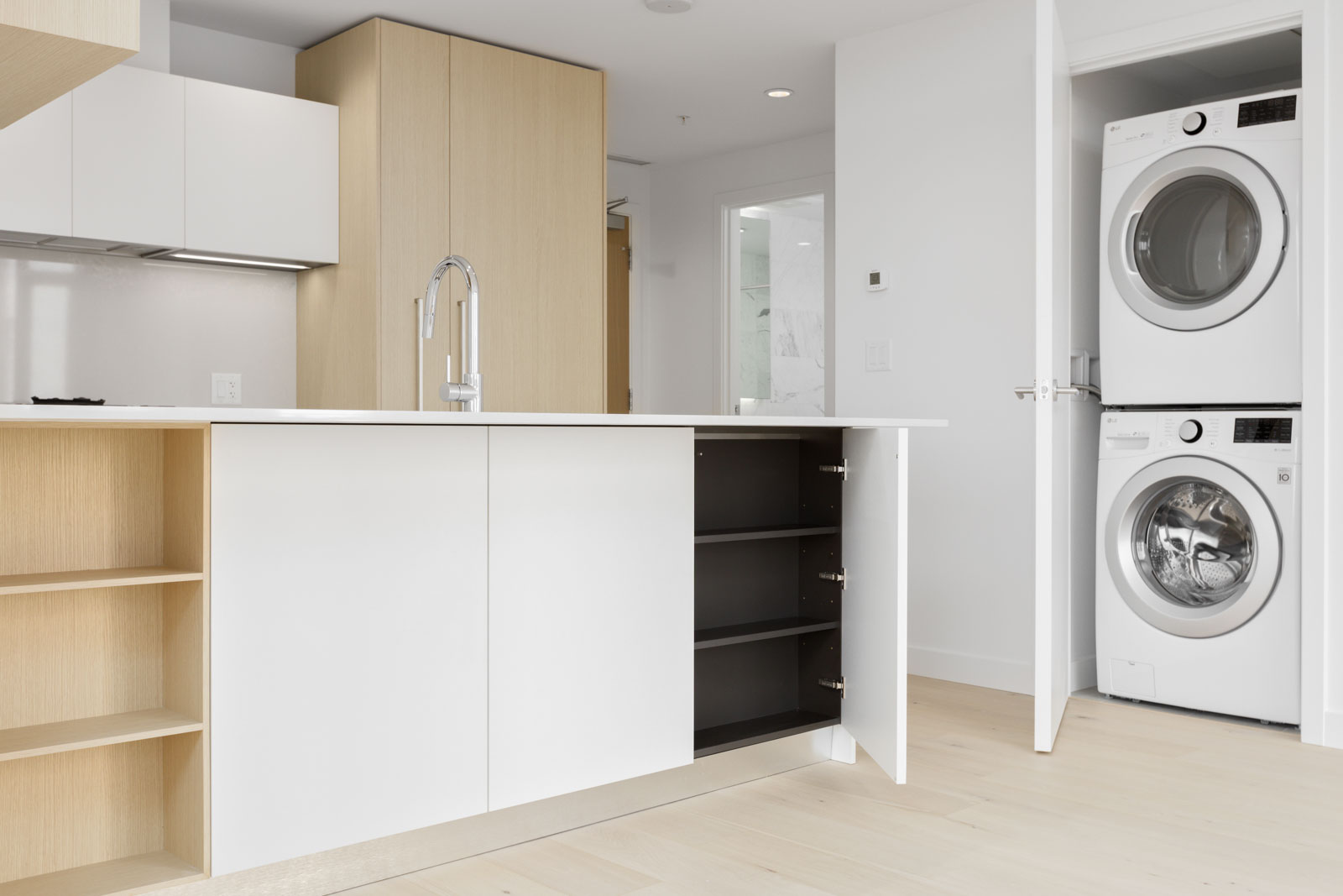 Modern kitchen with light wood and white cabinets, an island with open shelves, and a view of a stacked washer and dryer in an adjacent open closet.