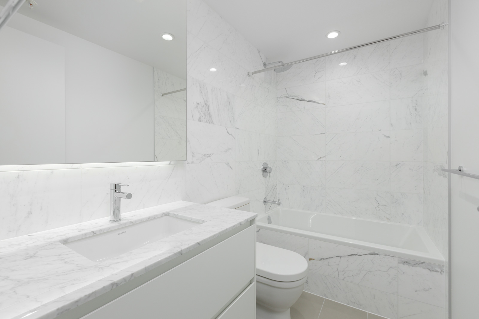 Modern bathroom with white marble surfaces, a bathtub with shower, a toilet, and a vanity with a sink and mirror under recessed lighting.