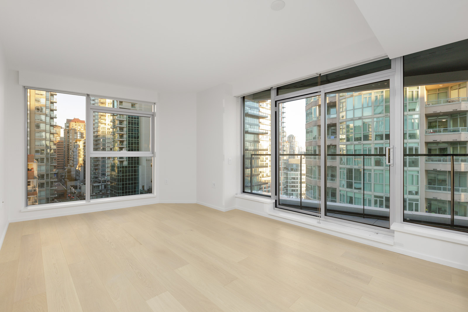 Bright, empty modern apartment room with large windows and sliding glass doors, light wood floors, and views of surrounding high-rise buildings.