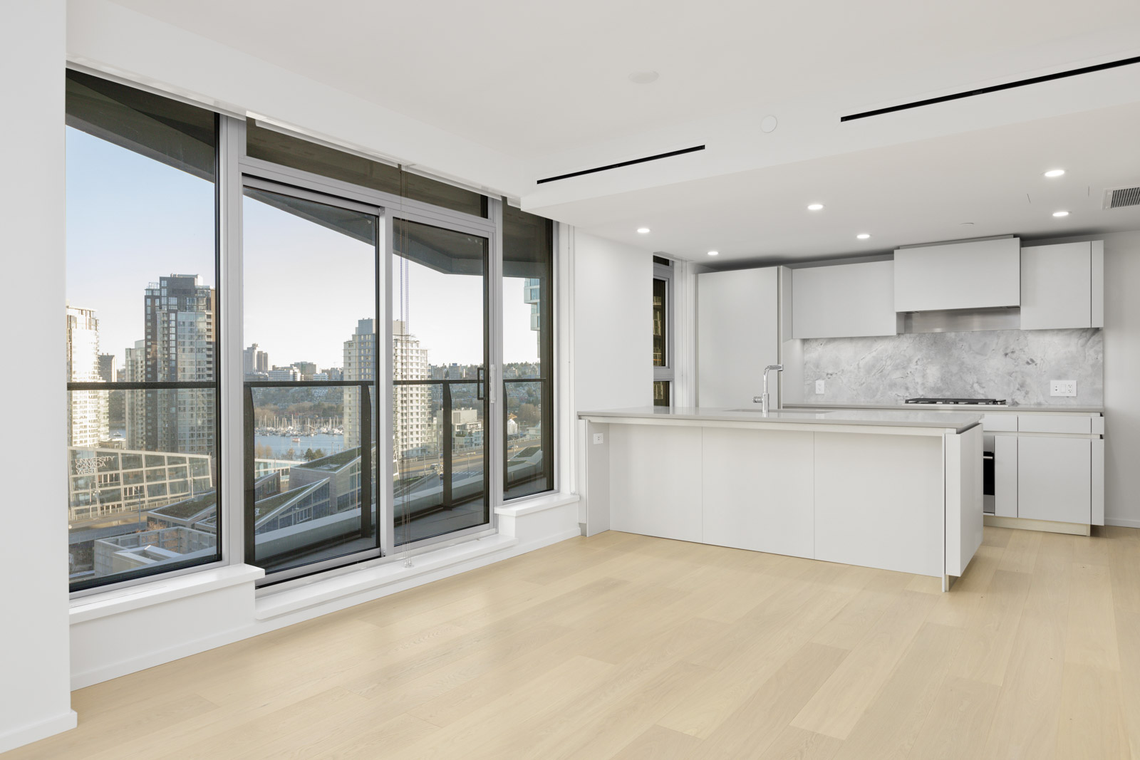 Modern, unfurnished apartment with large floor-to-ceiling windows, light wood floors, and an open kitchen featuring white cabinetry and marble backsplash. Cityscape visible outside.