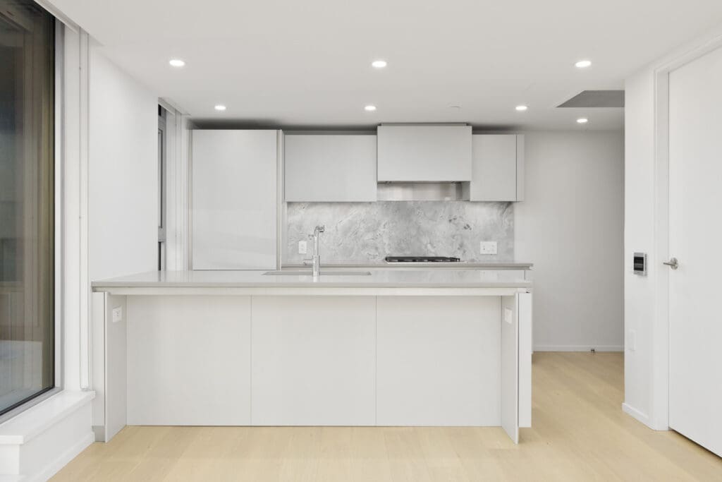 Modern kitchen with white cabinets, gray marble backsplash, built-in stovetop, and central island, illuminated by recessed ceiling lights and natural light from a large window.