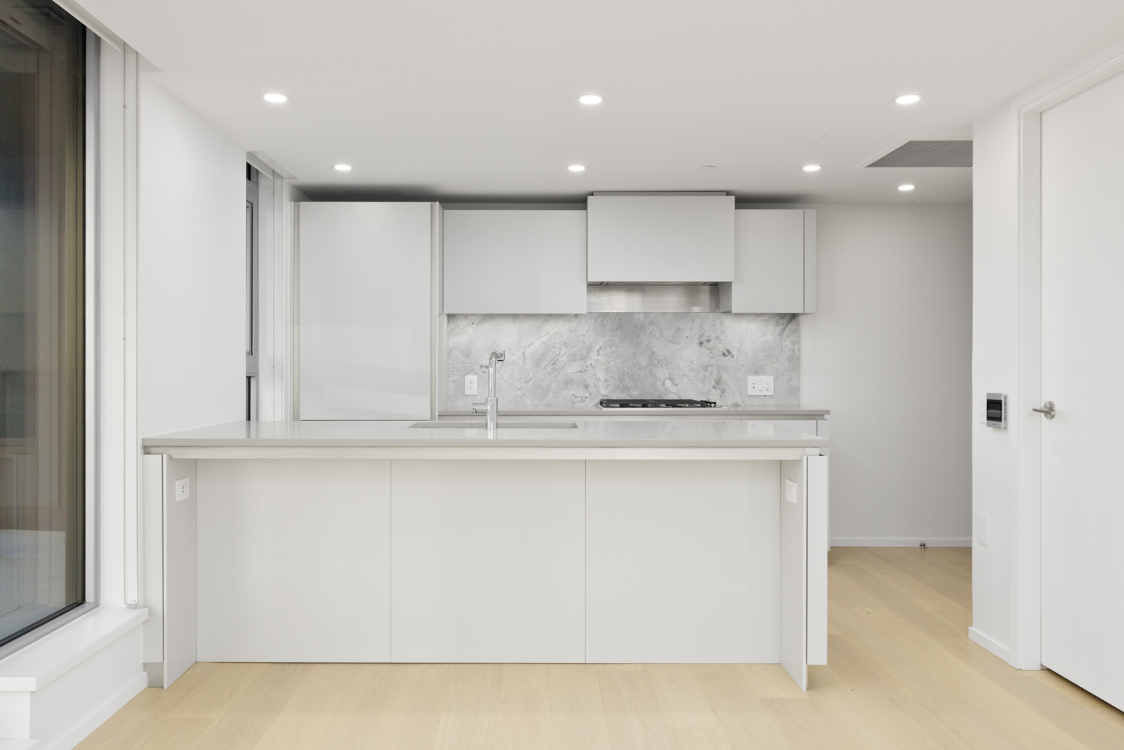 Modern kitchen with white cabinets, gray marble backsplash, built-in stovetop, and central island, illuminated by recessed ceiling lights and natural light from a large window.