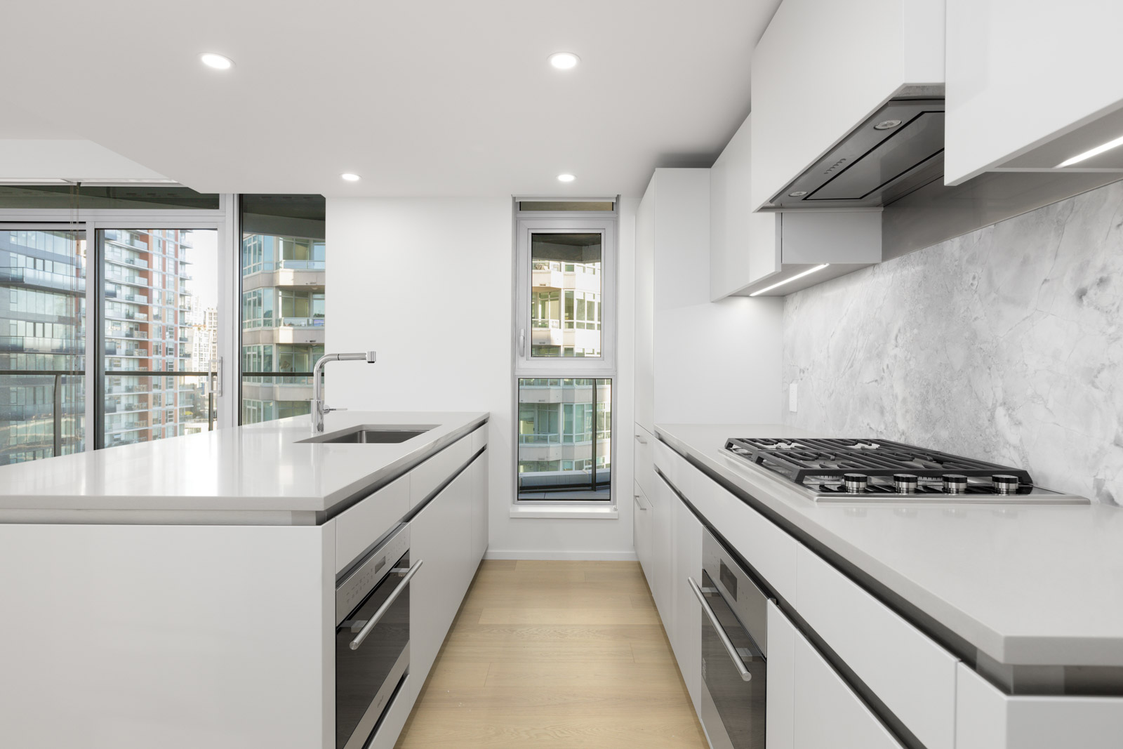 Modern kitchen with white cabinetry, built-in oven, gas cooktop, large island, sink, and floor-to-ceiling windows overlooking a cityscape.