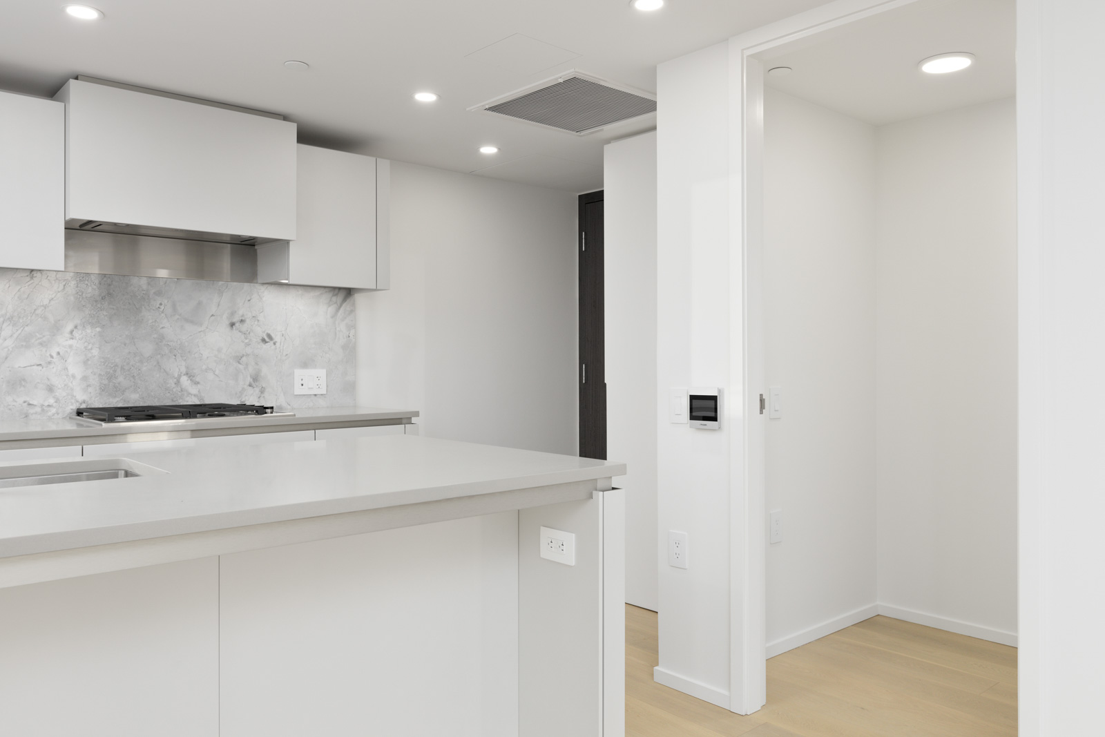 Modern kitchen with white cabinets, marble backsplash, light wood flooring, and an adjacent empty room with recessed lighting.