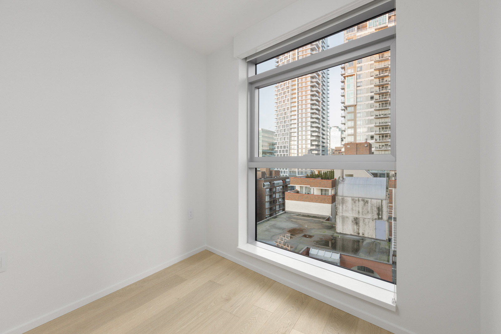 Empty white room with light wood flooring and a large window overlooking city buildings and rooftops on a clear day.