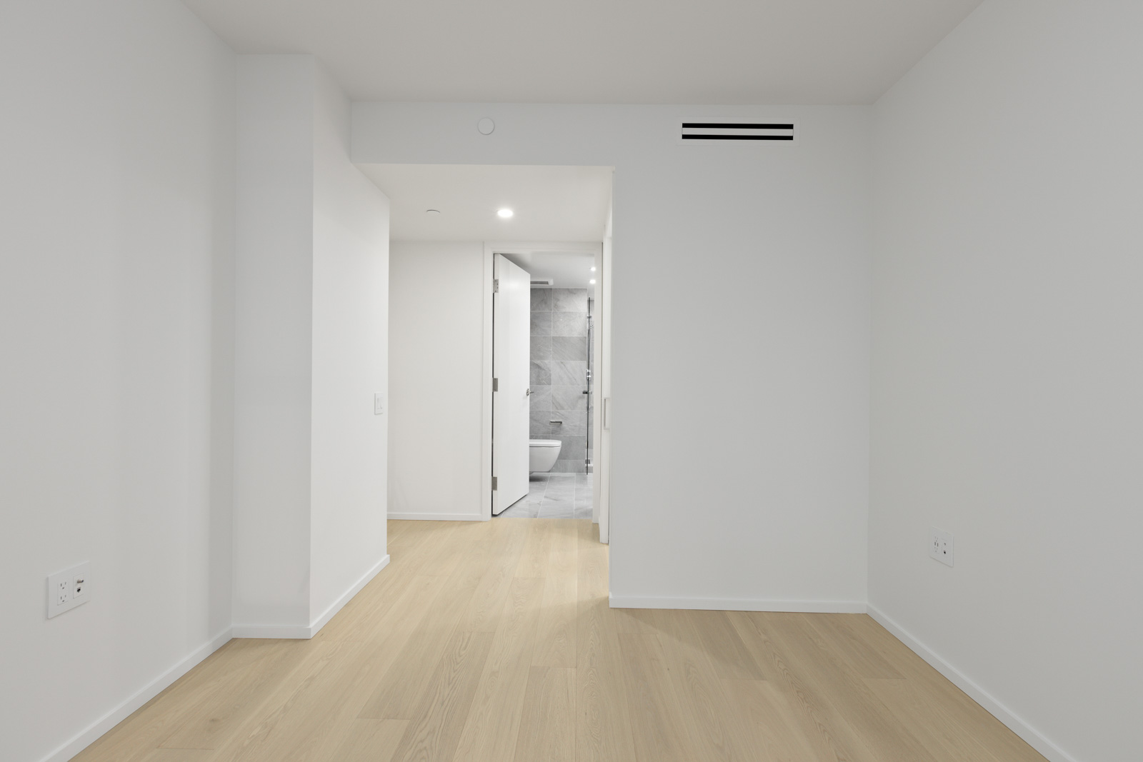 An empty room with light wood floors and white walls, facing a doorway that leads to a bathroom with gray tiles and modern fixtures.