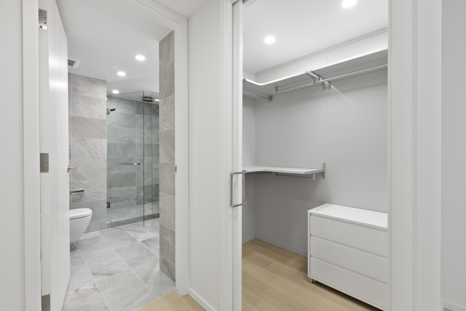 A walk-in closet with white shelves and drawers leads to a modern bathroom with gray tiles, a glass shower, and a toilet.