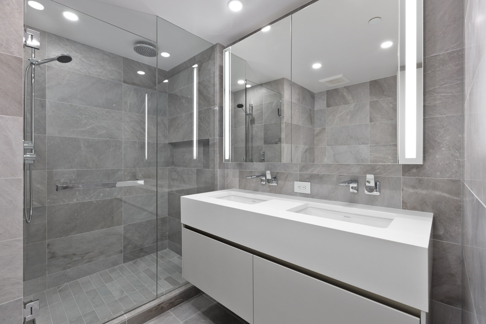 Modern bathroom with gray tile walls, a glass-enclosed shower, and a double sink vanity with large mirrors and integrated lighting.