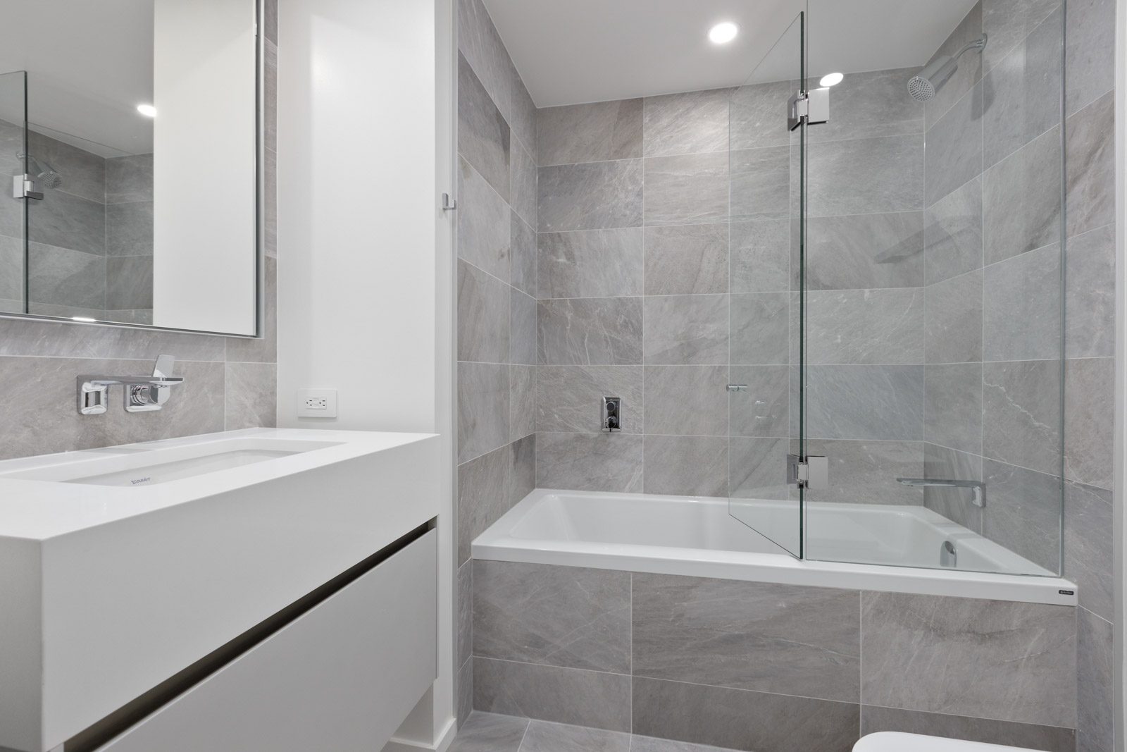 Modern bathroom with a white sink, wall-mounted faucet, large mirror, and a bathtub-shower combo with gray tile and glass partition.
