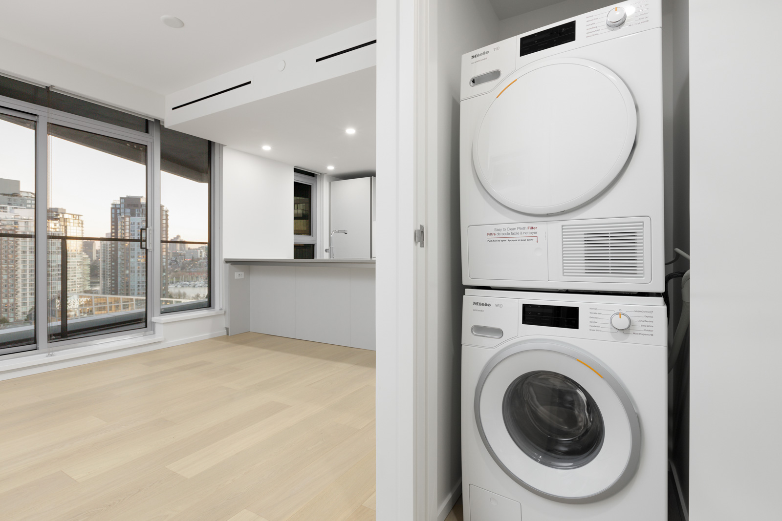 A stacked washer and dryer unit in a white closet next to a modern open-plan living area with large windows and light wood flooring.