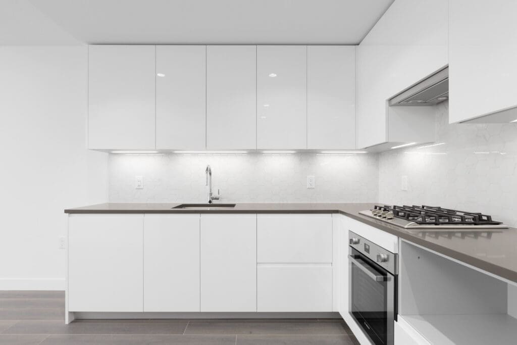 Modern kitchen with white cabinets, gray countertop, built-in oven, gas stovetop, under-cabinet lighting, and a single sink against a white hexagonal tile backsplash.
