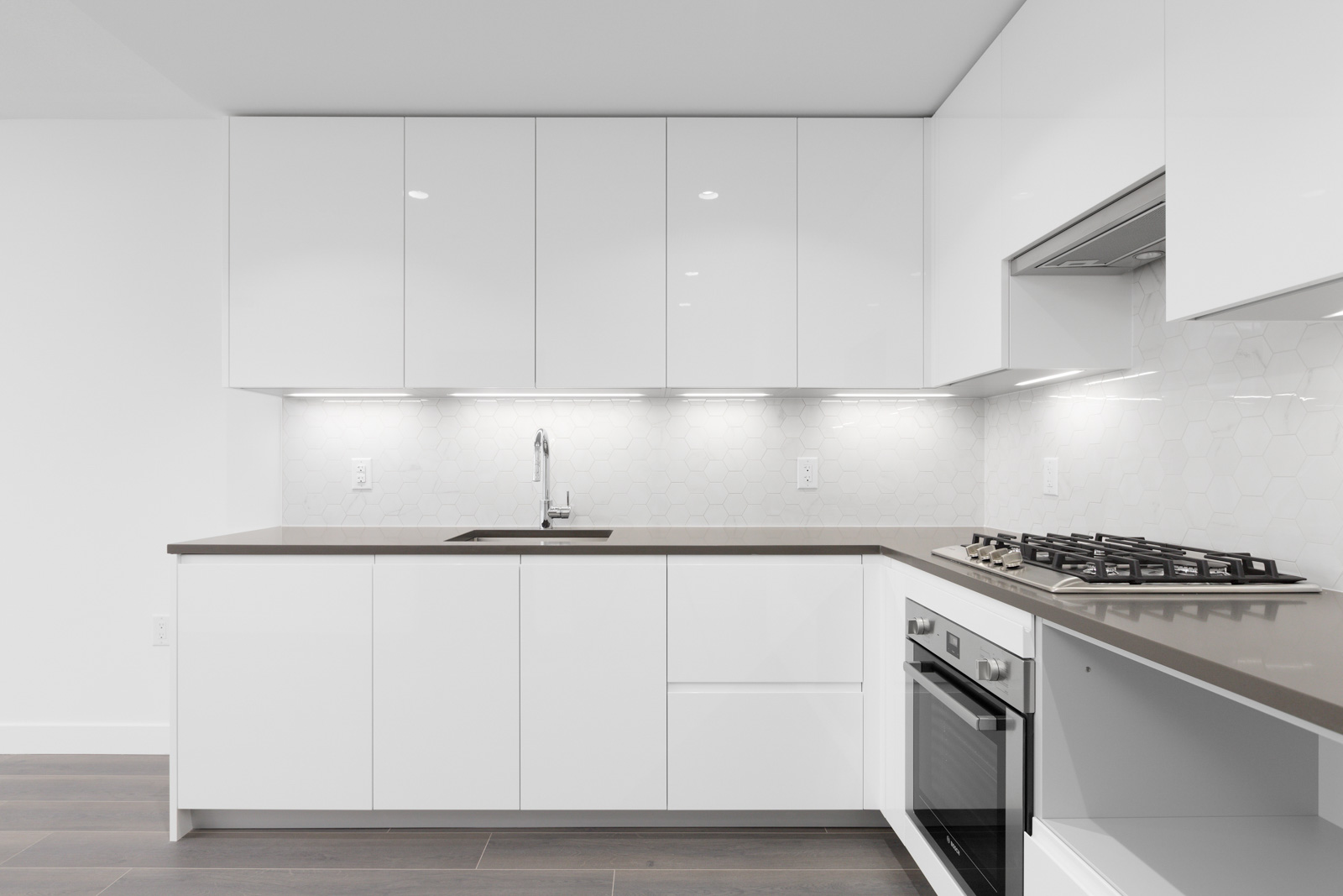 Modern kitchen with white cabinets, gray countertop, built-in oven, gas stovetop, under-cabinet lighting, and a single sink against a white hexagonal tile backsplash.