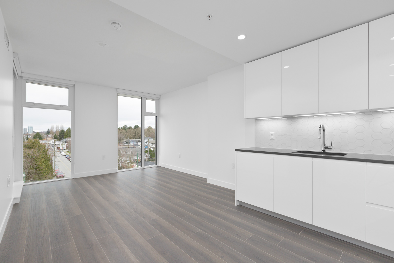 Unfurnished modern apartment with large windows, white cabinets, built-in kitchen sink, and dark wood flooring. Urban view visible through windows.