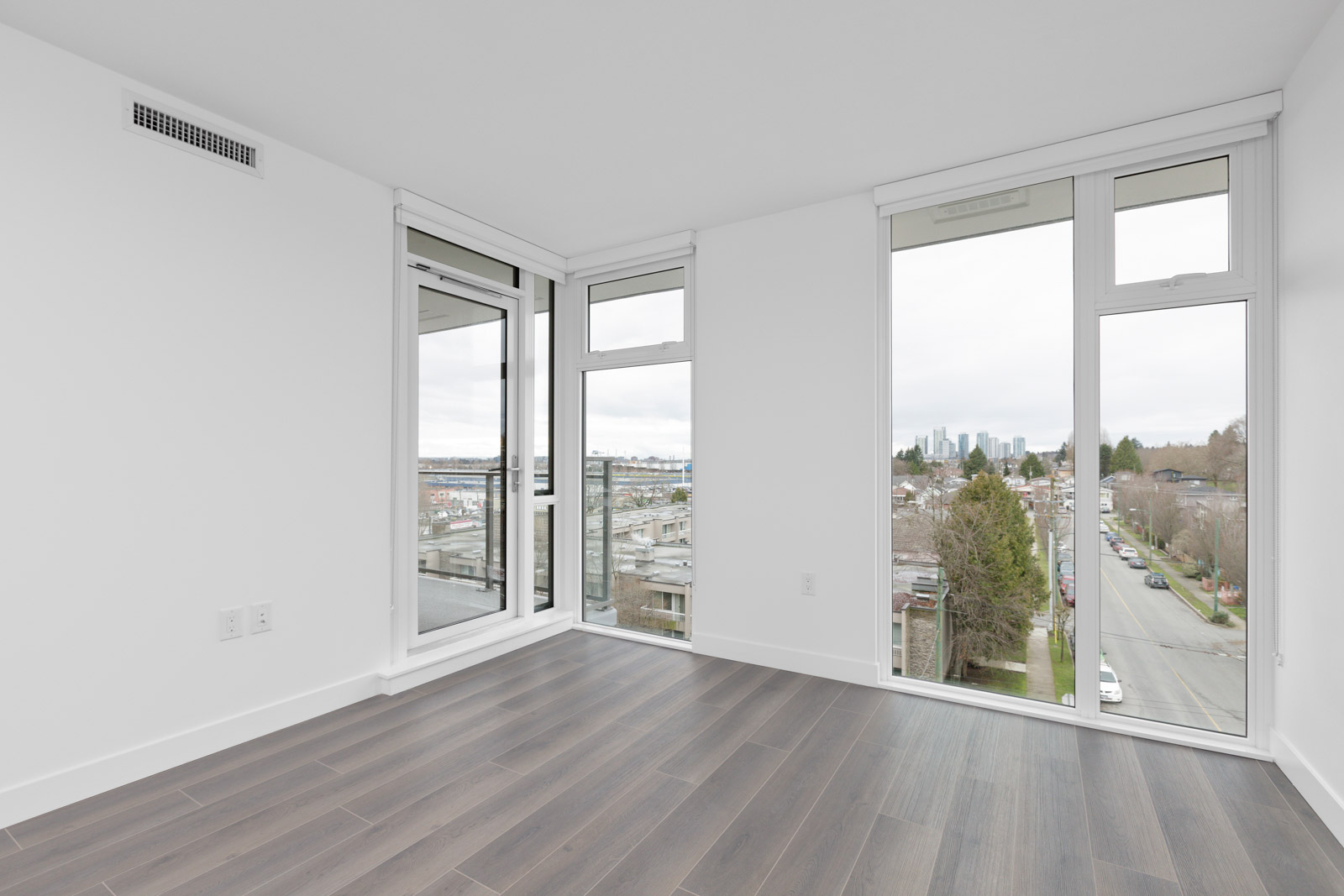 Empty modern apartment room with large floor-to-ceiling windows, wood flooring, and a view of a city street and skyline.