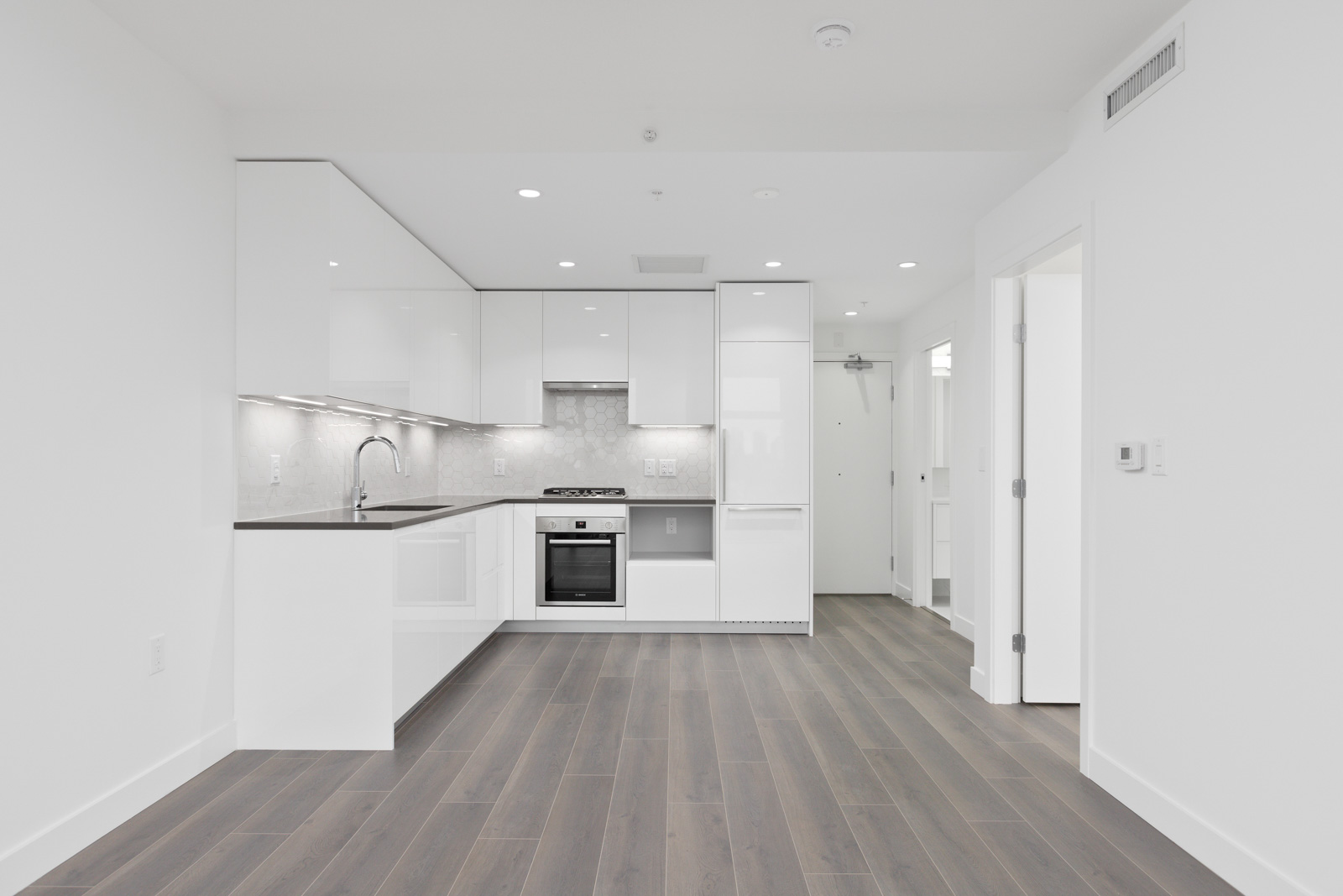 Modern, unfurnished white kitchen with built-in appliances, light gray backsplash, and dark wood flooring, adjacent to an open doorway and a hallway.